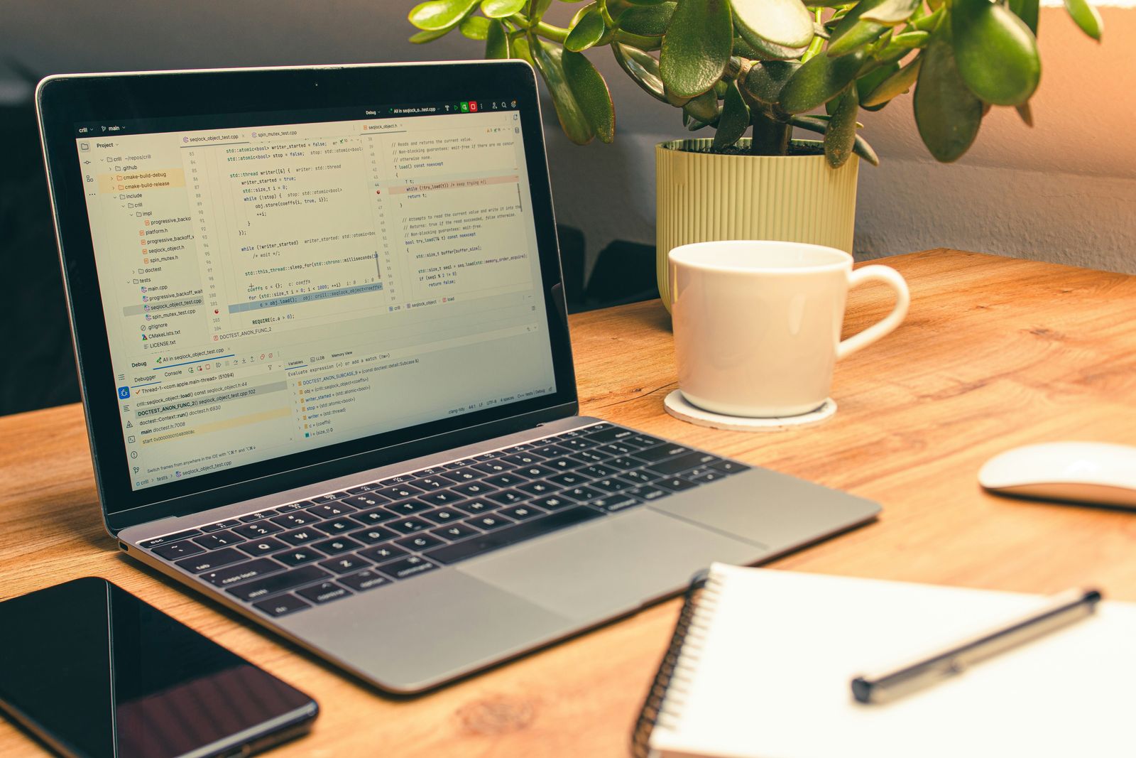 Laptop showing a code editor on a wooden desk beside a coffee mug, a potted plant and a notebook